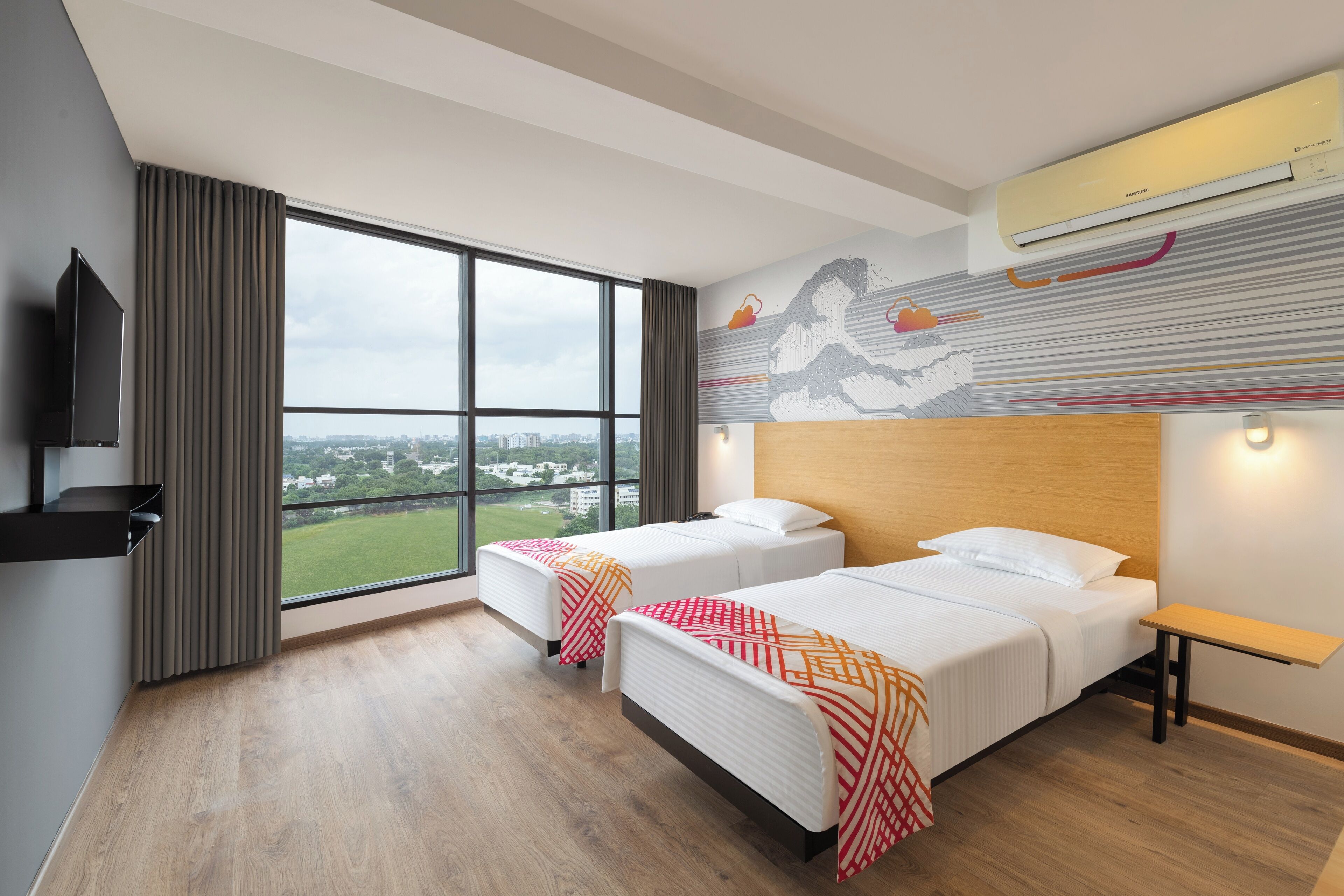 A hotel room with two beds, a large window with a city view, an air conditioning unit, and a mounted television.