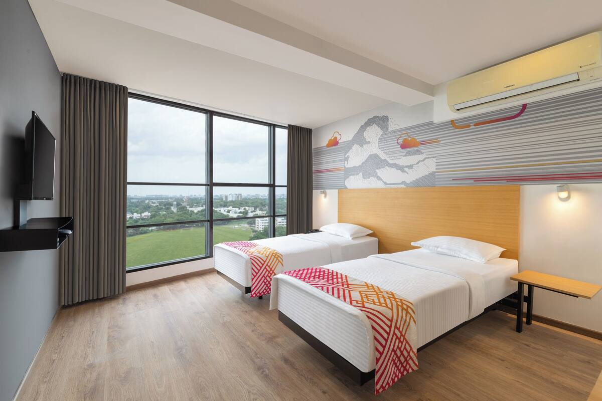 A hotel room with two beds, a large window with a city view, an air conditioning unit, and a mounted television.