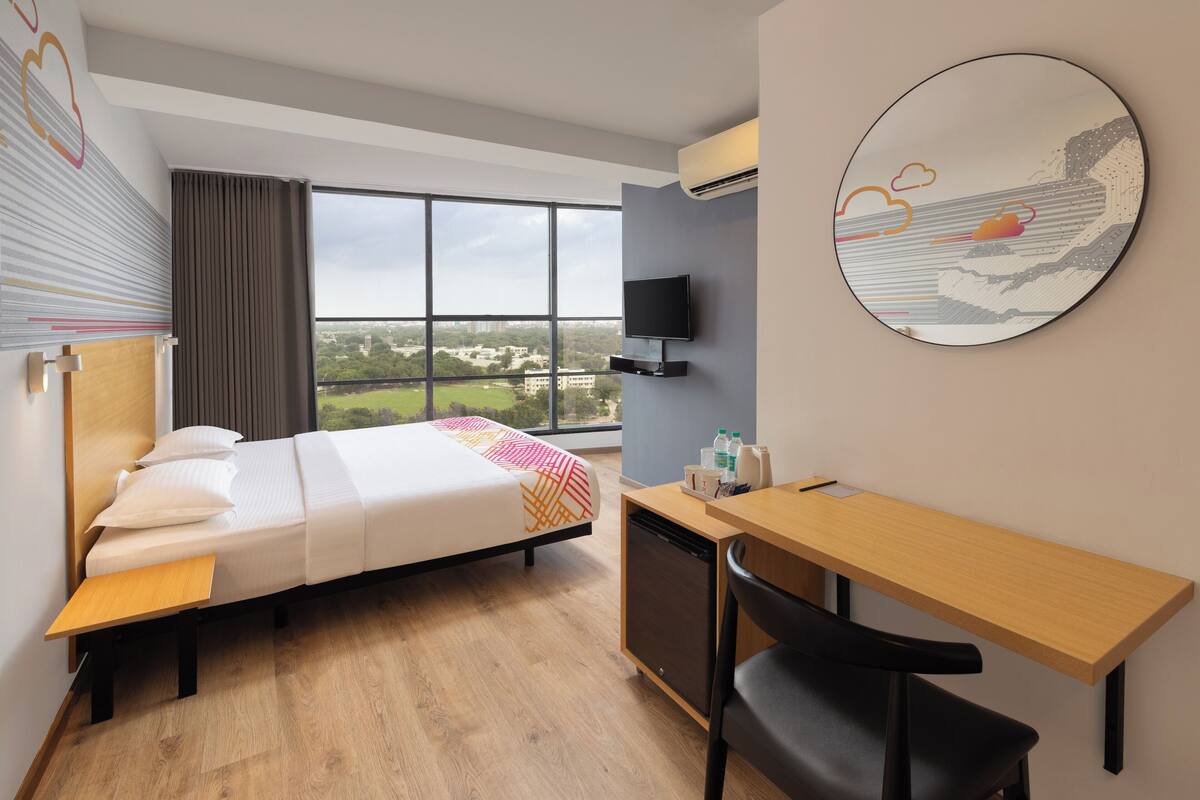 A hotel room with a bed, desk, chair, TV, and a large window with a city view.