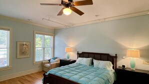 Une chambre avec un grand lit, un ventilateur de plafond, deux tables de chevet avec des lampes, un banc et une fenêtre avec des stores.