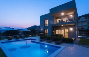 Une maison moderne à deux étages, dotée d’une piscine, d’un espace salon extérieur et offrant une vue sur les montagnes.
