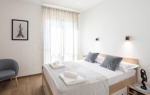 Un lit bien fait, avec du linge de lit blanc et gris, une chaise grise et, au mur, un tableau représentant la Tour Eiffel.