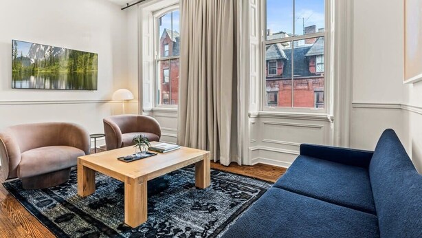 Un salon moderne avec une table basse en bois, deux fauteuils, un canapé et de grandes fenêtres avec des rideaux.