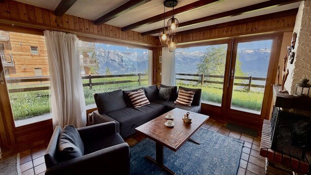 A cozy room with a sofa, a table with cups, and a view of mountains through large windows.