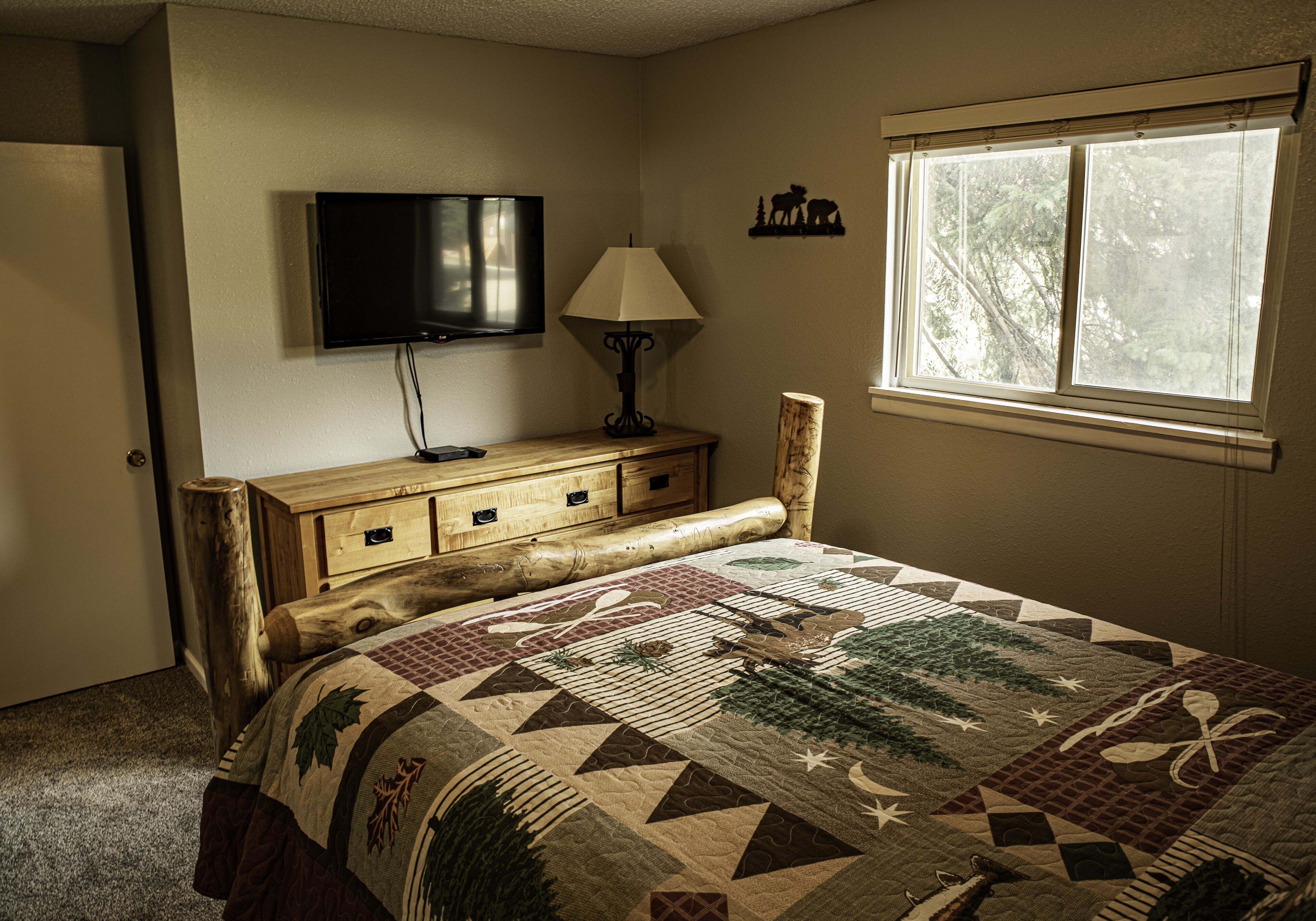 A bedroom with a wooden headboard, a television mounted on the wall, a bedside lamp, a window with a view of trees, and a quilt with a nature theme.