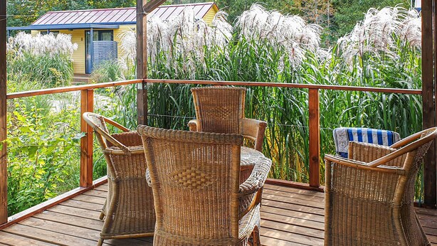 A wooden deck with wicker chairs and a view of tall grasses and a yellow building in the background. - Cozy home in Biskupiec with lake view (Biskupiec)