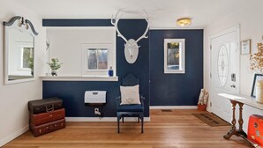 Een kamer met een donkerblauwe accentmuur, een houten vloer, een witte radiator, een blauwe stoel, een witte deur met een vluchtwijzer en een grote decoratie van een hertenschedel.