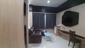 Ein modernes Wohnzimmer mit einem braunen Sofa, einem Glastisch, einem an der Wand befestigten Fernseher und einem dunklen Vorhang, der die Fenster verdeckt.