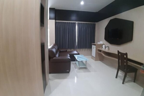 A modern living room with a brown sofa, a glass coffee table, a TV mounted on the wall, and a dark curtain covering the windows. - Tho Mai Resort by Zuzu (Khlong Yai)