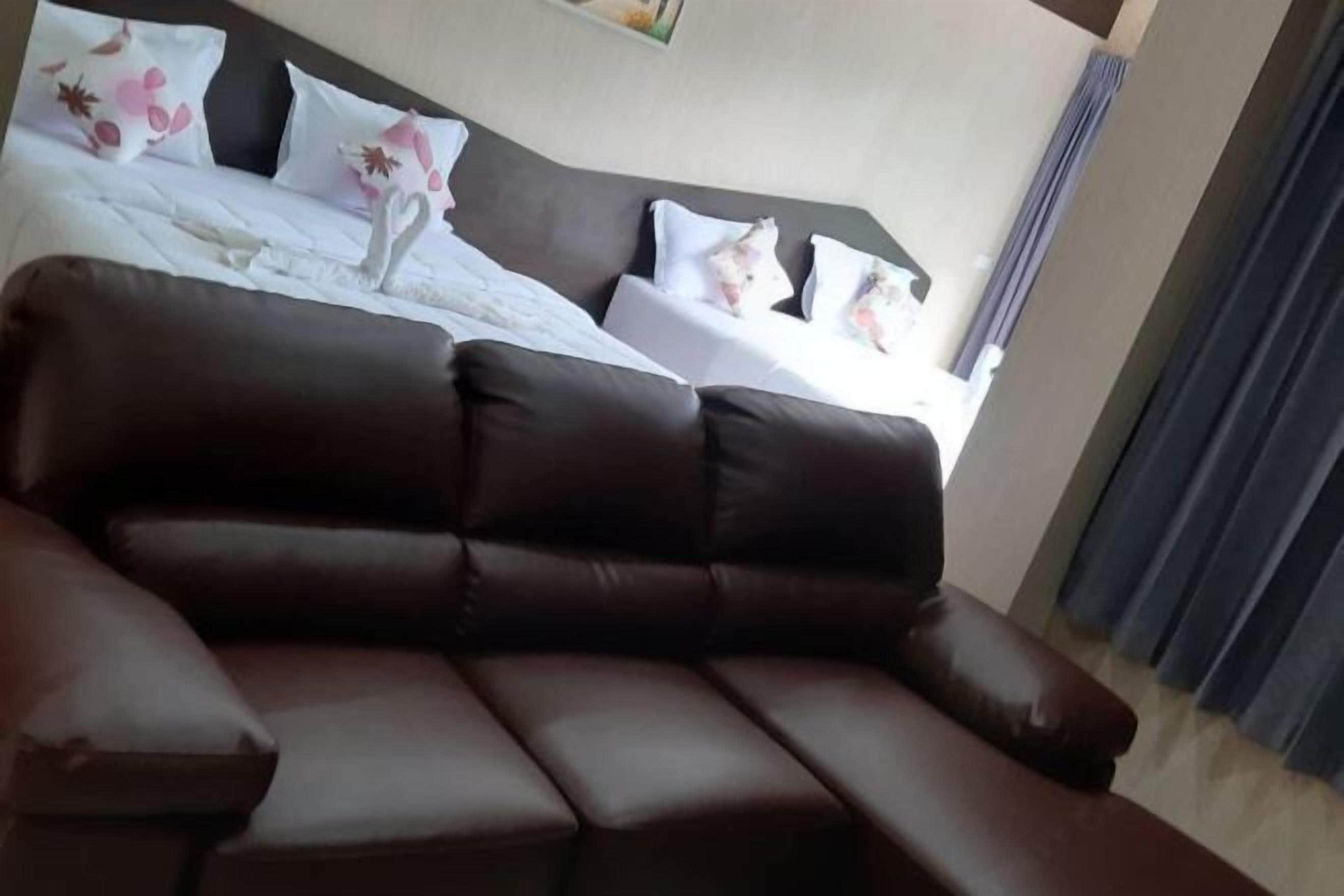 Habitación de hotel con sofá de cuero marrón oscuro, cama bien hecha con almohadas estampadas con motivos florales y un amplio ventanal con cortinas.