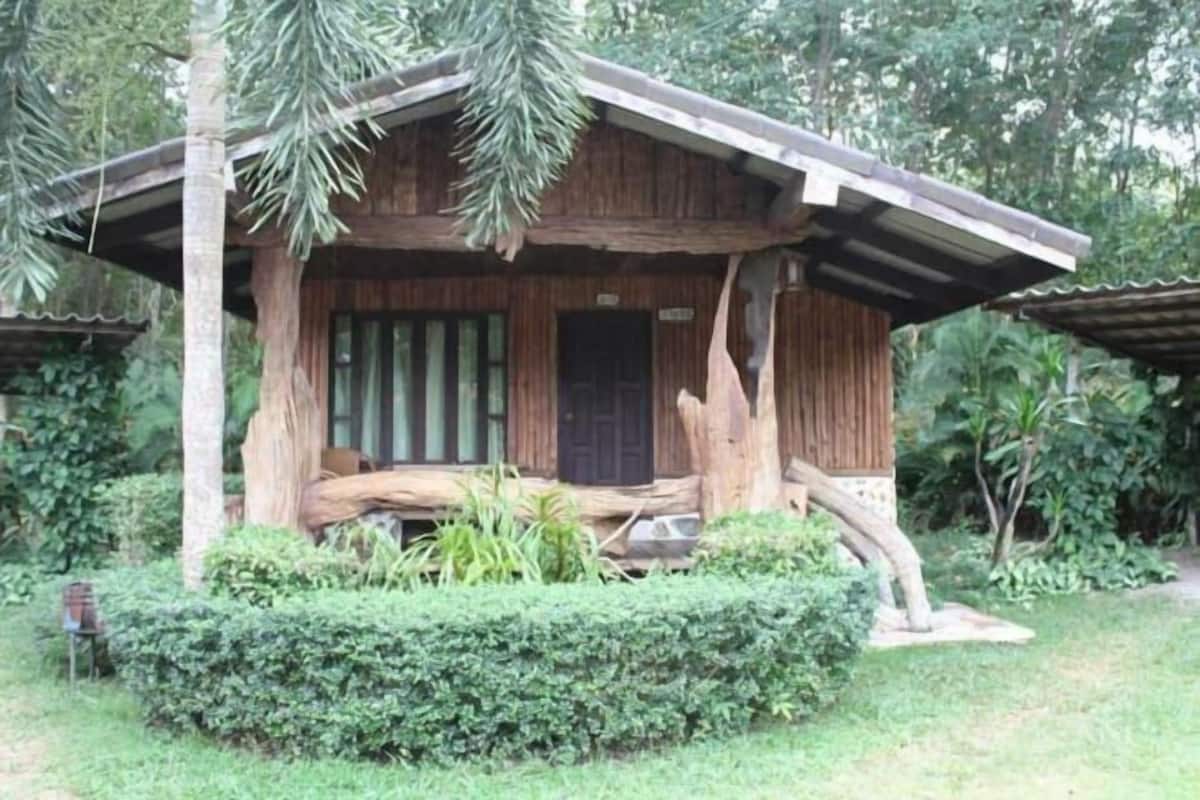 A rustic wooden cabin with a porch, surrounded by lush greenery.