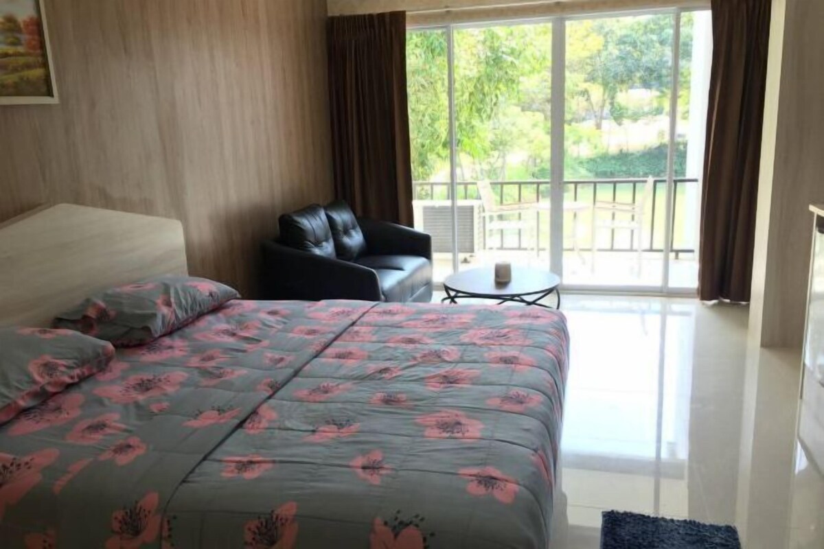 A bedroom with a bed, a chair, a small table, and a balcony with a view of greenery.