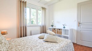 Un dormitorio con una cama grande, una mesita de noche, una lámpara, una ventana con cortinas, una pared blanca, una puerta y una estantería con cestas y un jarrón.