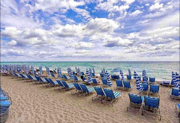 Una playa con filas de sillas azules y sombrillas a rayas, de frente al océano.