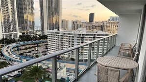 Un balcon avec vue sur le paysage urbain, comprenant des immeubles de grande hauteur et une route sinueuse. On y trouve des espaces de détente en plein air avec des tables et des chaises.