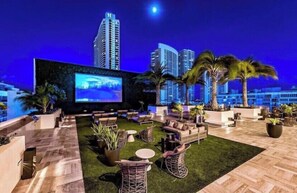 A rooftop lounge with outdoor seating, a large screen, and palm trees.