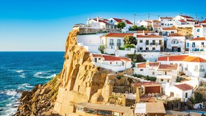 Uma vila costeira com edifícios brancos, telhados de terracota e um penhasco rochoso com vista para o mar.