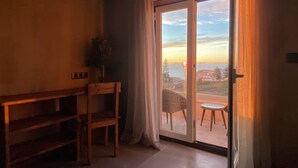 Quarto com um banco de madeira, uma cadeira e vista para o pôr do sol sobre a costa.