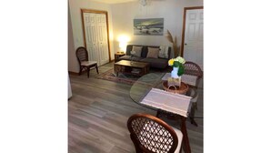 Un salon comprenant un canapé, une table basse et une table à manger avec des chaises. On y trouve également un ventilateur de plafond, un tableau au mur et une porte.