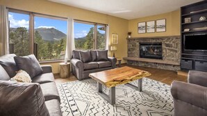 A living room with a stone fireplace, leather sofas, a coffee table, and large windows offering a mountain view. - Exceptional Views, hot tub -A Mountain Life! #3245 (Estes Park)
