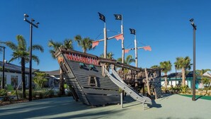 A pirate-themed play structure with slides and climbing areas, surrounded by palm trees and a clear sky.