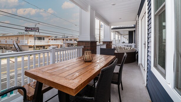 A covered balcony with a wooden table and wicker chairs, overlooking a street with buildings and a sign reading "Coral Sands". - Spacious Beach Block Home: 3 Beds/2 Baths, Front Porch w/ Furniture Included (Ocean City)
