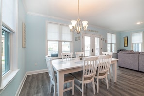 A dining area with a white table and chairs, a chandelier, and a view of the beach through the windows. - Spacious Beach Block Home: 3 Beds/2 Baths, Front Porch w/ Furniture Included (Ocean City)