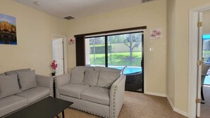Ein Wohnzimmer mit zwei grauen Sofas, einem schwarzen Couchtisch, einem Bild von einem See und einer Tür, die auf eine Terrasse mit Blick auf eine Rasenfläche und einen Swimmingpool führt.