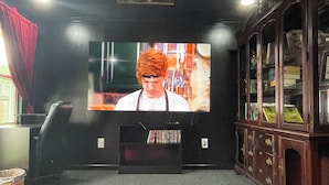 Ein Zimmer mit an der Wand montiertem Fernseher, einem Bücherregal und roten Kissen auf dem Boden.