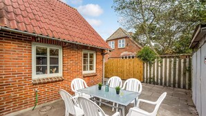 Eine Terrasse mit Glastisch und weißen Stühlen, ein Ziegelhaus mit roten Dachziegeln und ein Holzzaun.