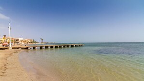 Una spiaggia con un molo di legno che si protende verso il mare, acque limpide e poco profonde, e edifici sullo sfondo.