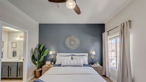 Un dormitorio con cama, mesitas de noche, ventilador de techo, un adorno en la pared y una ventana con cortinas.
