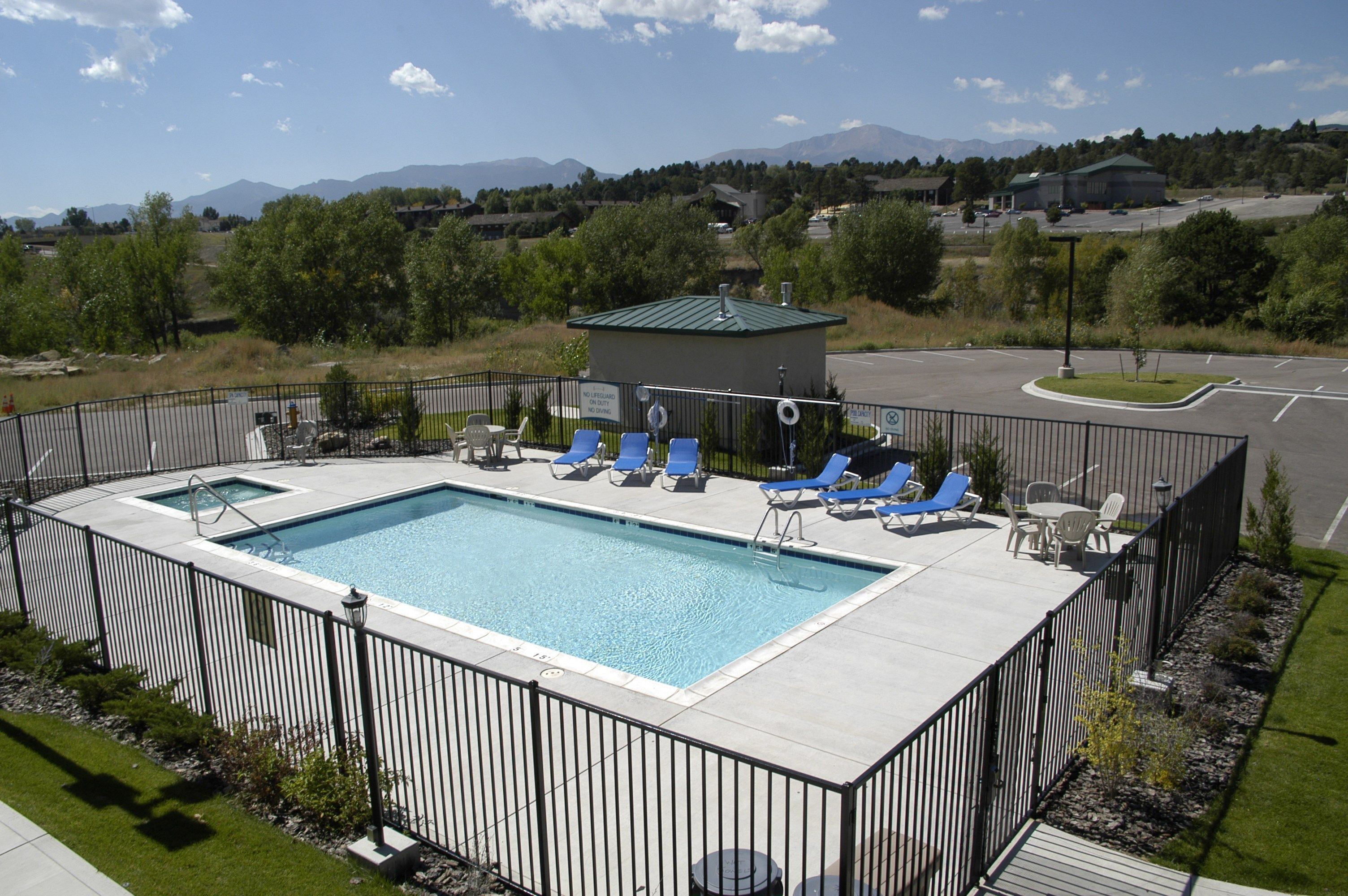 A rectangular swimming pool with a fenced deck, surrounded by blue lounge chairs and a small building with a green roof.