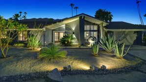 Een modern huis met een goed verlichte oprit, omringd door diverse planten en palmbomen.