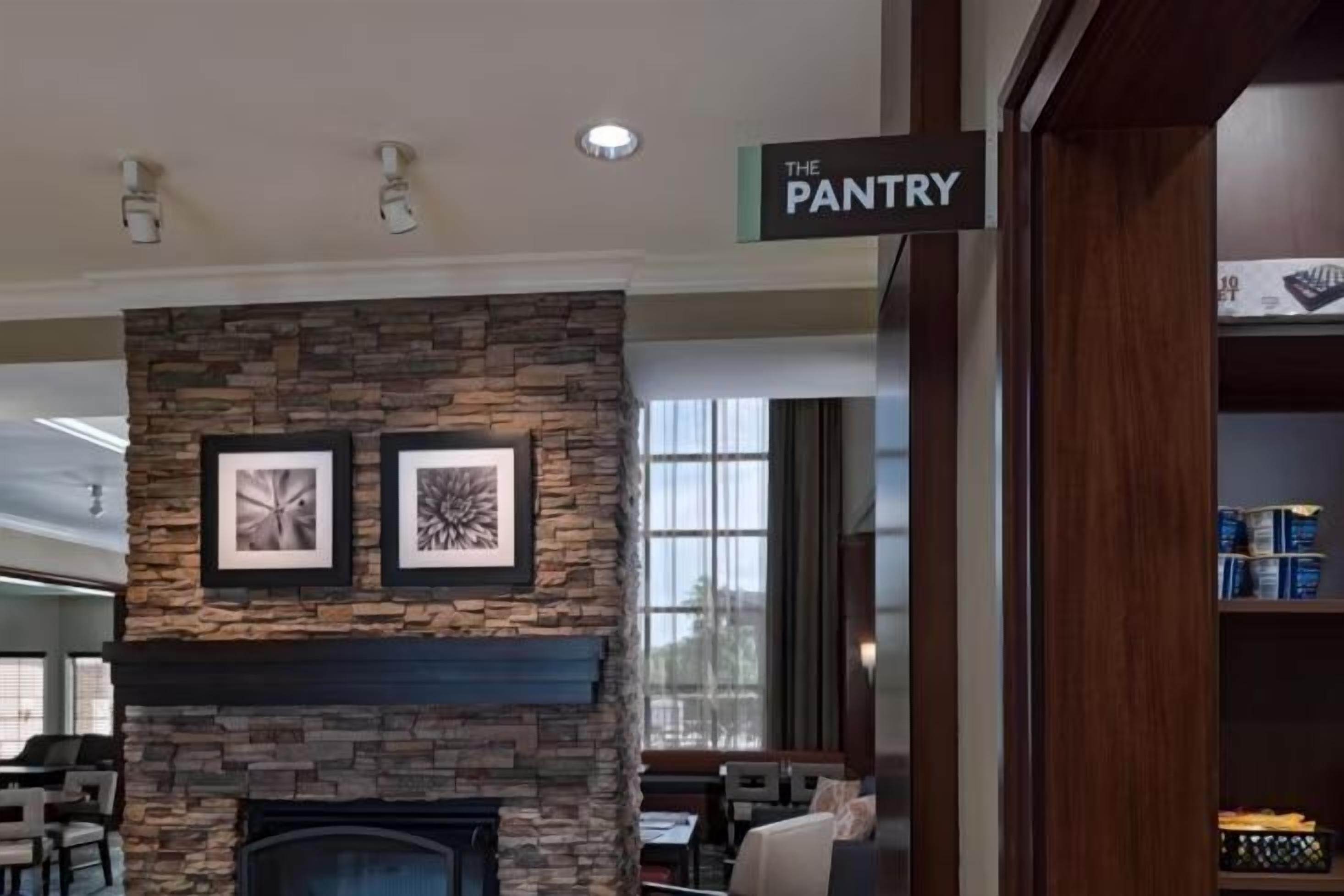 Una chimenea de piedra con obras de arte enmarcadas, un cartel que dice 'THE PANTRY', y vistas al exterior a través de amplios ventanales.