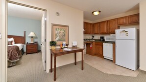Quarto de hotel com cama, mesa de cabeceira, abajur, quadro emoldurado e uma pequena mesa com uma planta e folhetos. O quarto possui piso coberto de carpete e uma porta que dá para o banheiro.