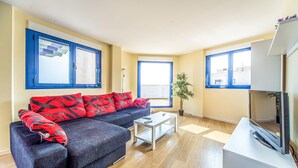 Un salon moderne avec un canapé gris foncé, des coussins à motifs rouges, une table basse blanche et un téléviseur à écran plat posé sur un meuble blanc.