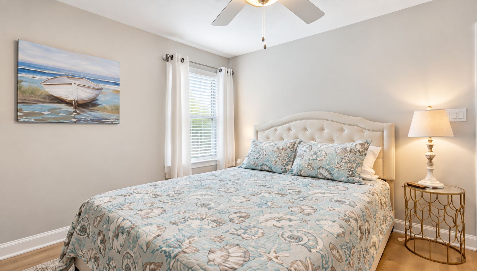 A bedroom with a bed, a nightstand, a ceiling fan, a window with curtains, and a painting on the wall.