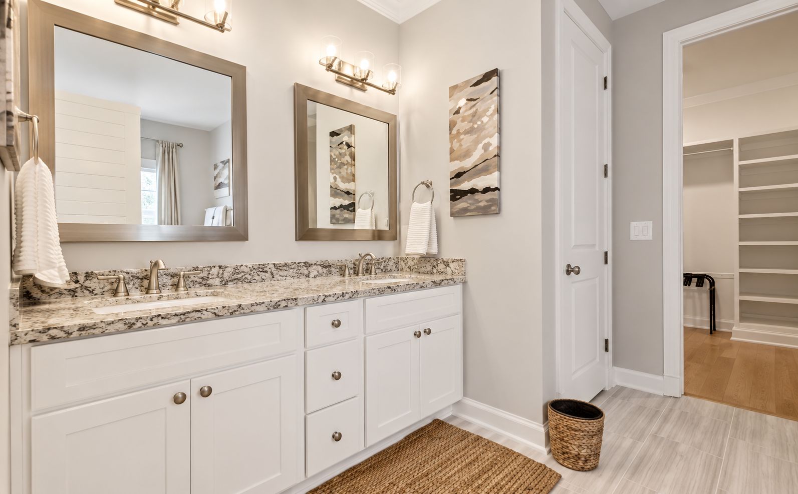 A bathroom with a double vanity, a large mirror, a wall-mounted light fixture, and a woven mat on the floor.