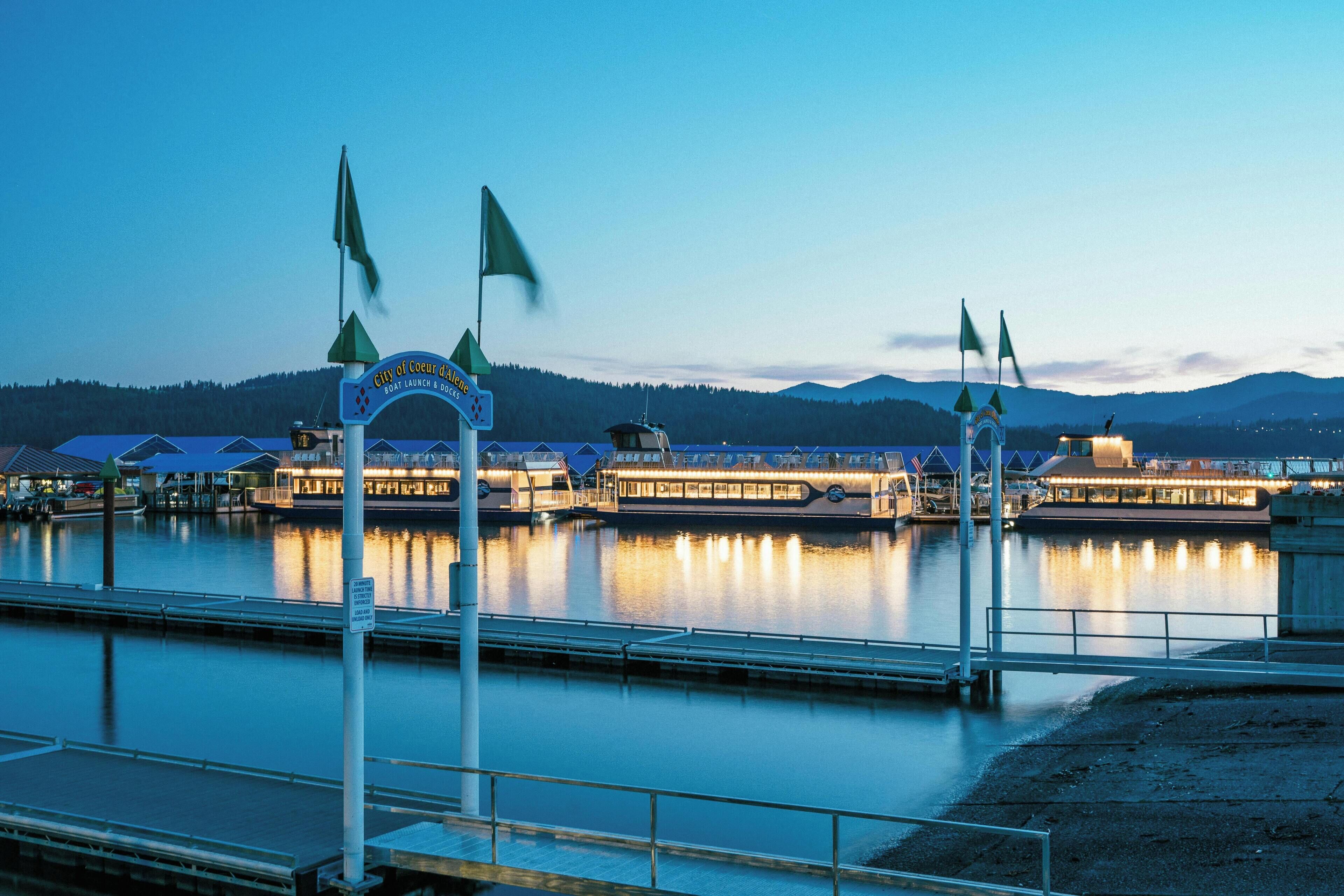 A waterfront hotel with a dock, a sign reading "City of Coeur d'Alene," and a view of the mountains in the background.