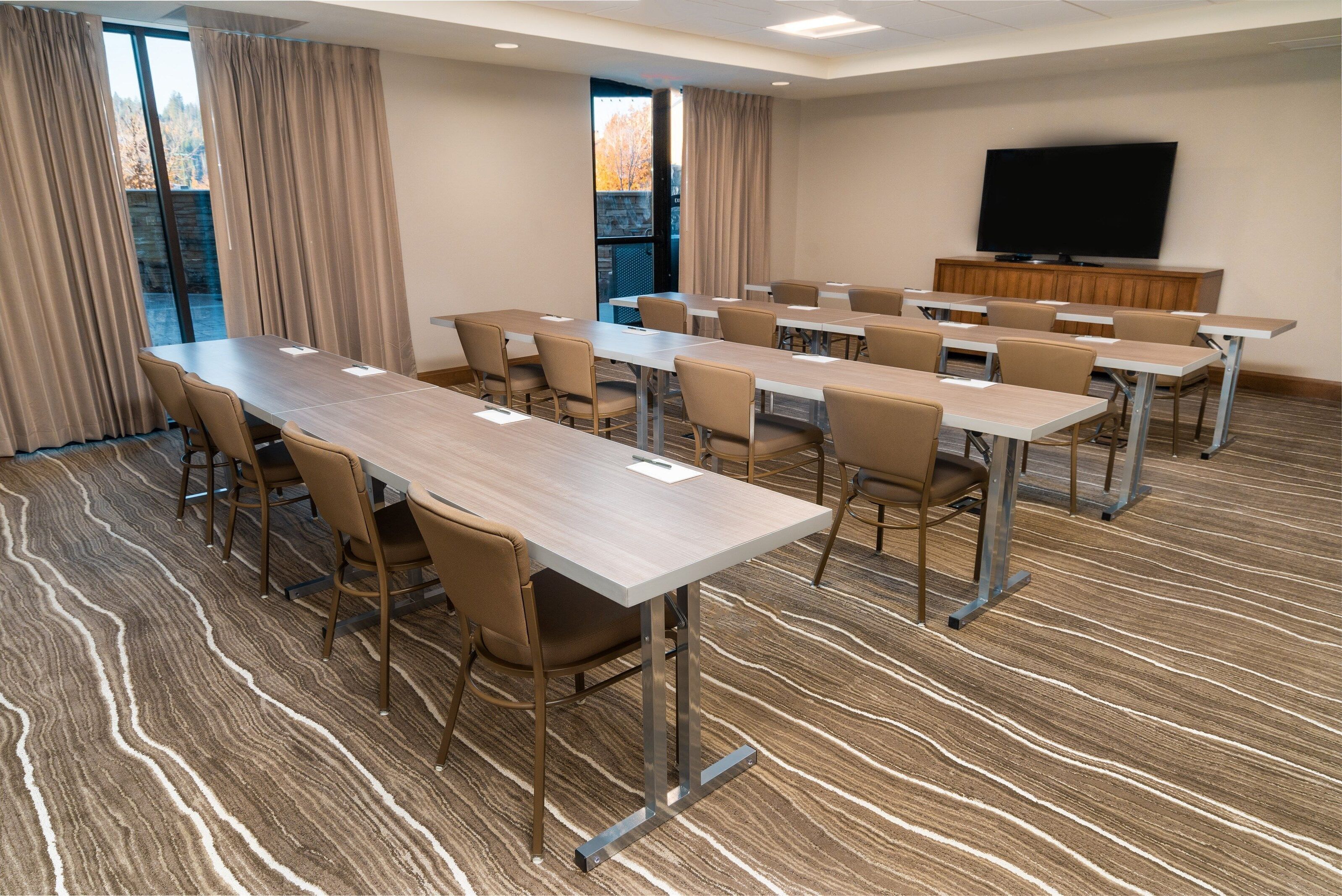 A conference room with a long table, chairs, a television, and a view of trees through large windows.