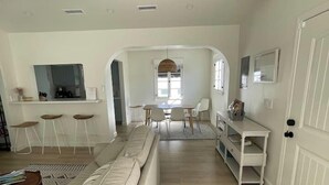 Un grand espace de vie comprenant un canapé, une table à manger et des chaises, un coin cuisine avec un comptoir et des tabourets, et un couloir menant à une porte.