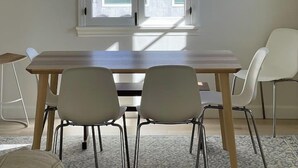 Un coin repas comprenant une table en bois, deux chaises, un luminaire suspendu en fibres tressées et une fenêtre avec des stores.