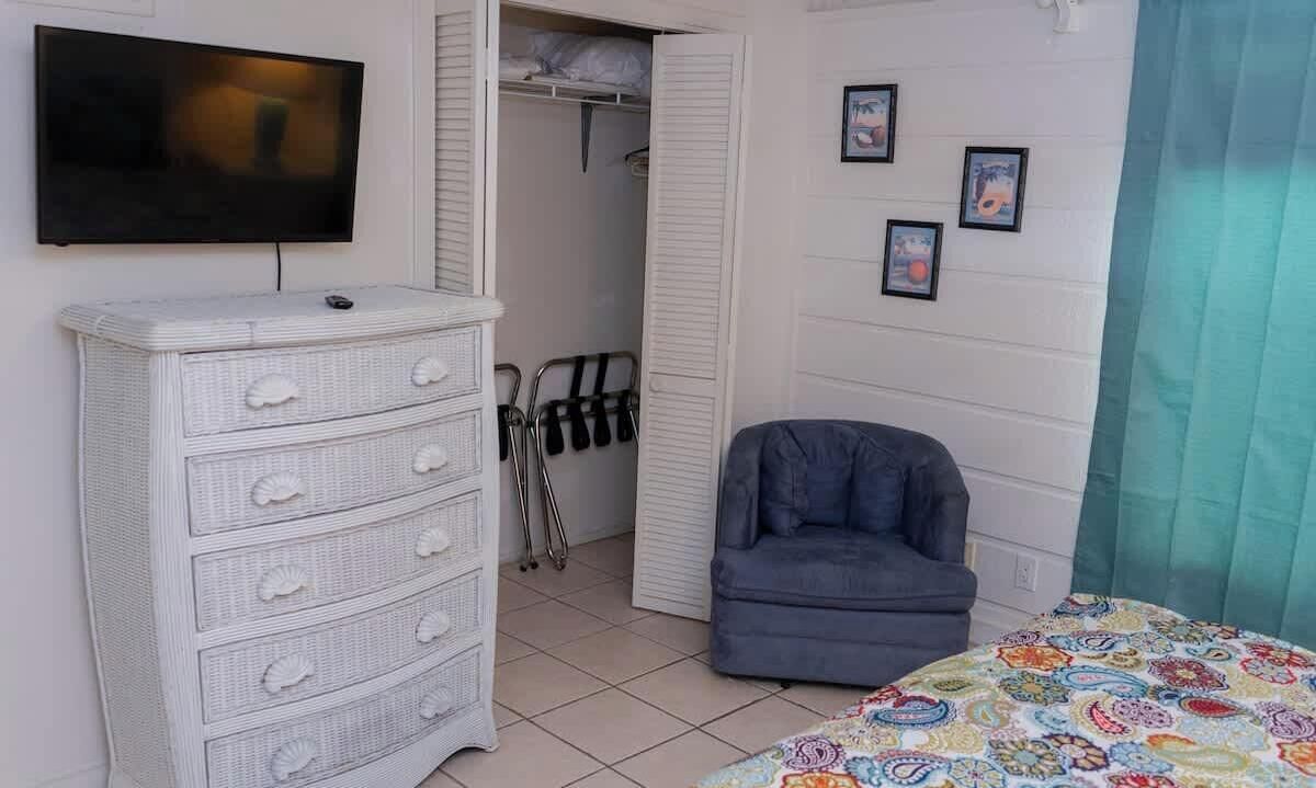A bedroom with a TV, a white dresser, a blue chair, and a bed with a colorful blanket.