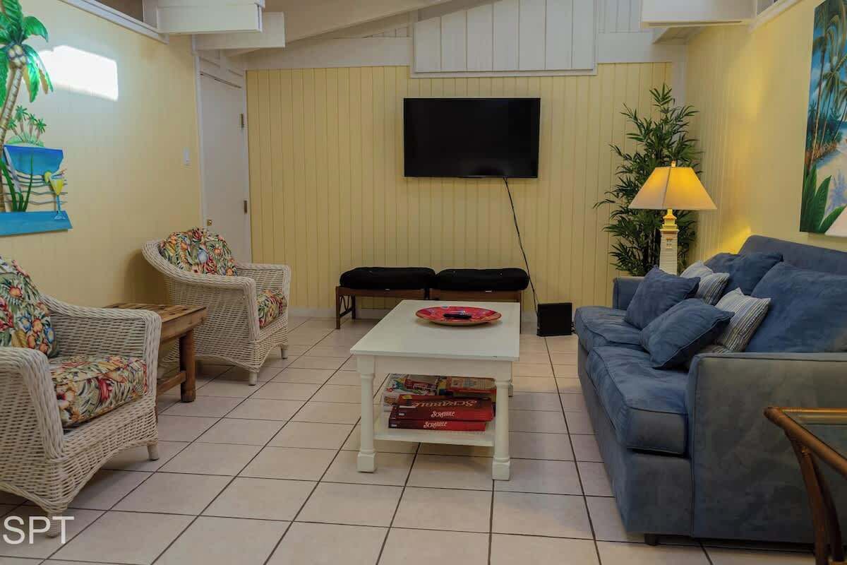 A living room with wicker furniture, a white coffee table with books, a blue sofa, and a TV on the wall.
