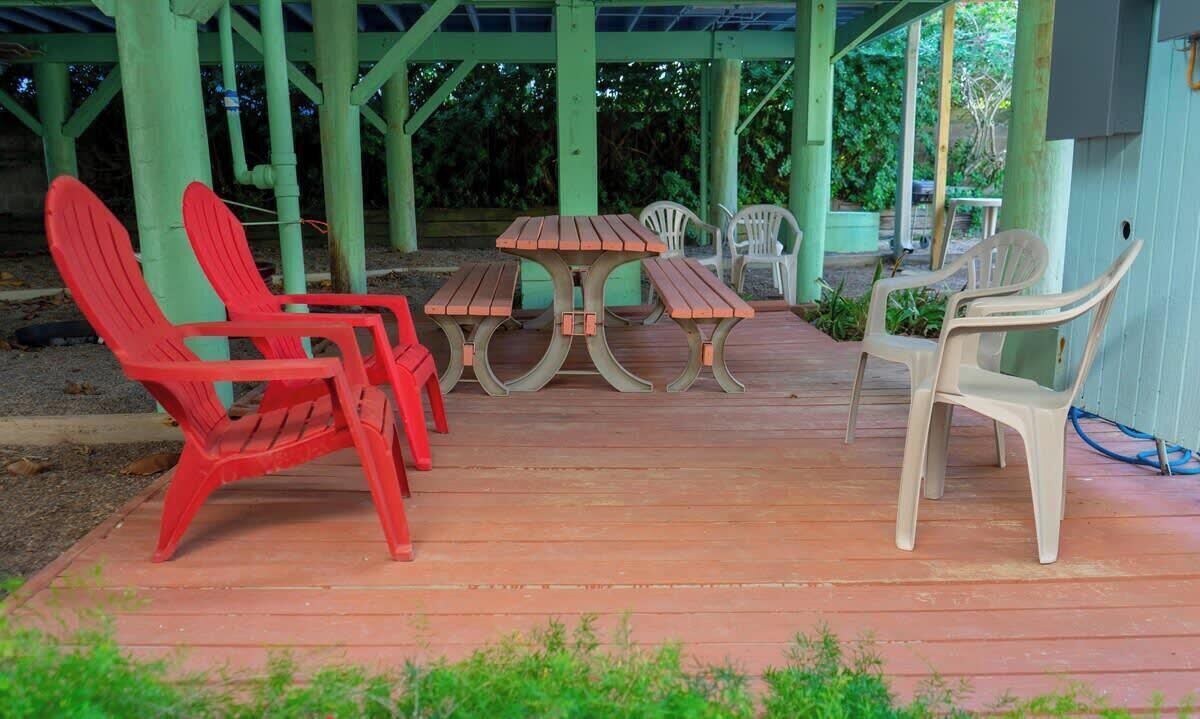 Une terrasse extérieure avec une table en bois et des bancs, entourée de piliers verts et de chaises en plastique.