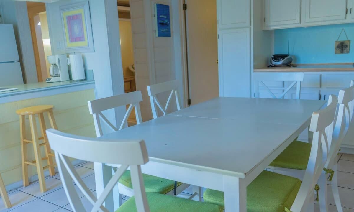 A dining area with a white table and chairs, a wooden stool, and a kitchen area with cabinets and appliances.