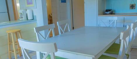 A dining area with a white table and chairs, a wooden stool, and a kitchen area with cabinets and appliances.