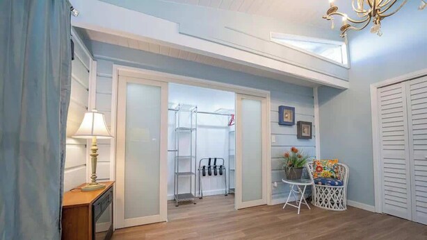 A room with a wooden floor, a small table with a lamp, a chair, a white wardrobe, and a glass door leading to a balcony with a clothes drying rack. - Warm Vibes - 5 Mins to Beach - Cool Retro Feel (South Padre Island)