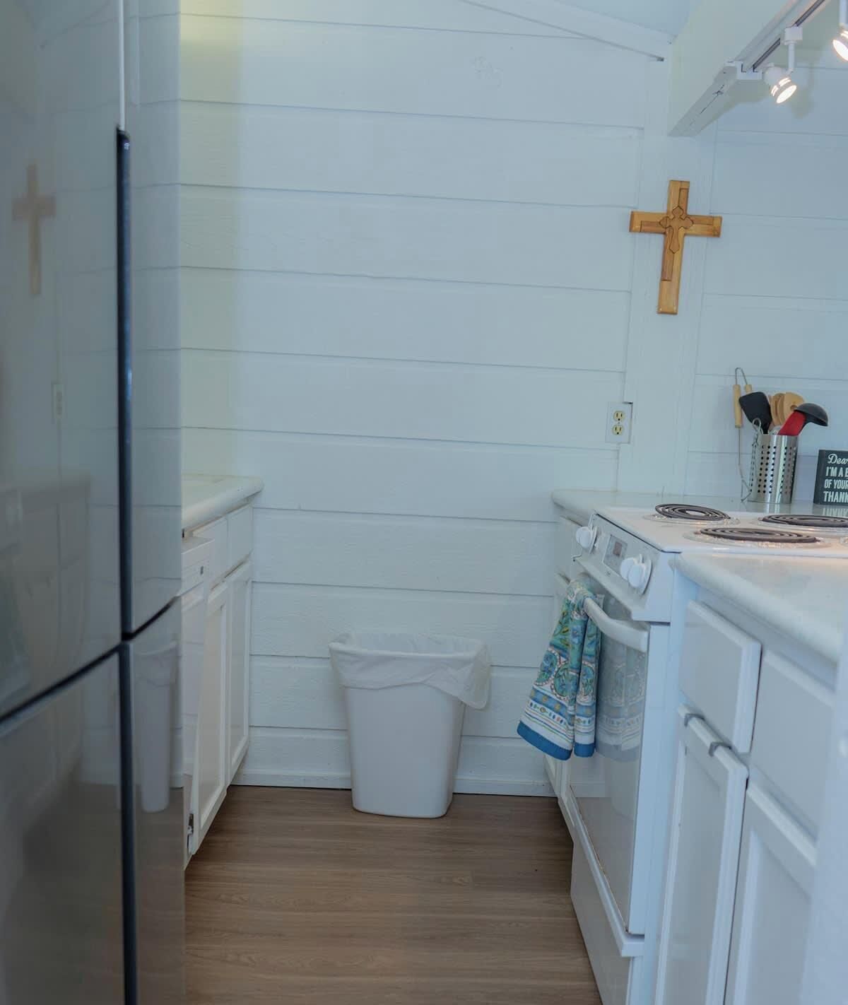 Une cuisine de taille réduite avec des meubles blancs, une croix en bois accrochée au mur, une poubelle et une cuisinière sur laquelle est suspendue une serviette à la porte du four.